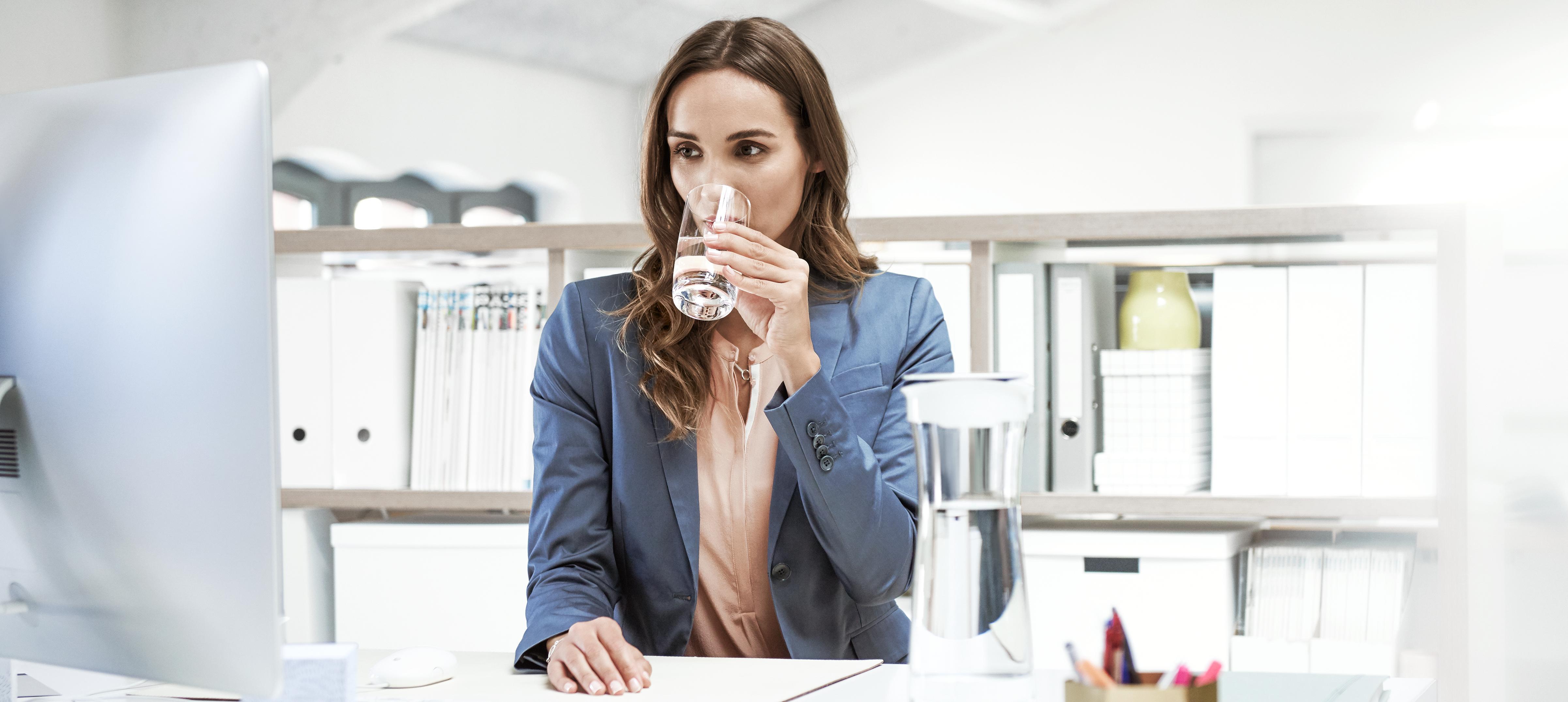 Kobieta pracująca w biurze, pijąca wodę ze szklanki podczas obowiązków służbowych.